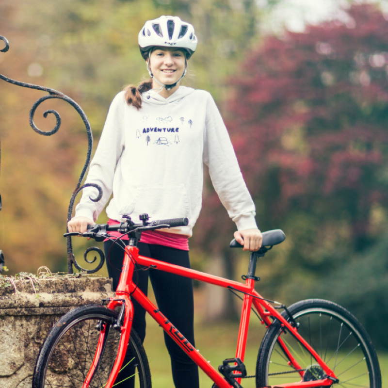 A girl wearing a helmet and holding a Frog 67 red bicycle in an outdoor setting