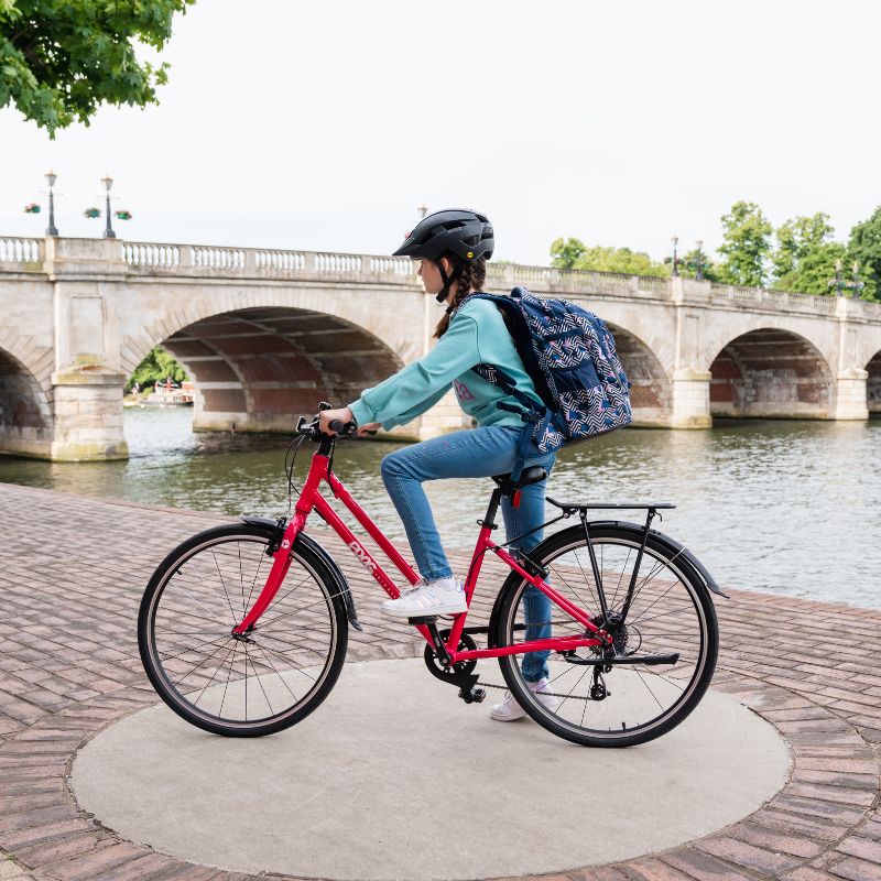 Kid riding a City 67 bicycle with a blue backpack near a river and bridge.