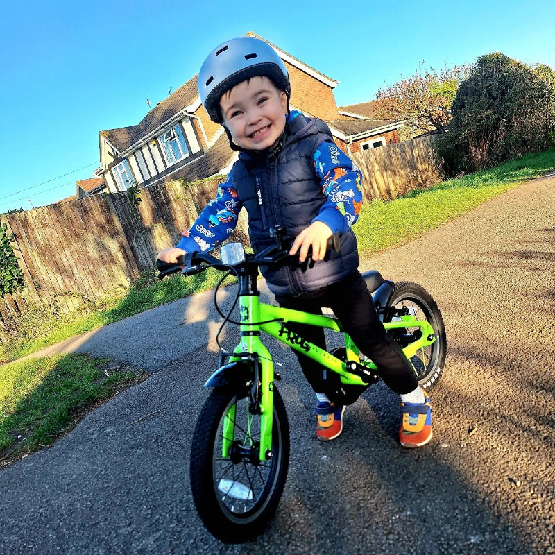 Child on a Frog 38 green bicycle wearing a helmet and colorful clothing in a residential area.