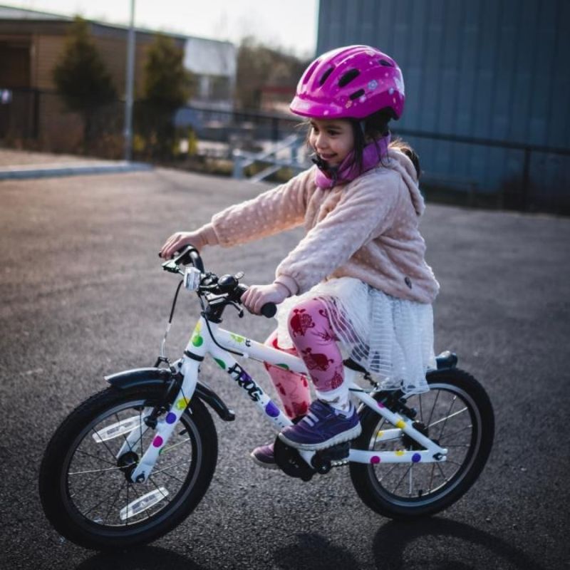 Child riding a Frog44 pedal bike with a pink helmet on an outdoor road.