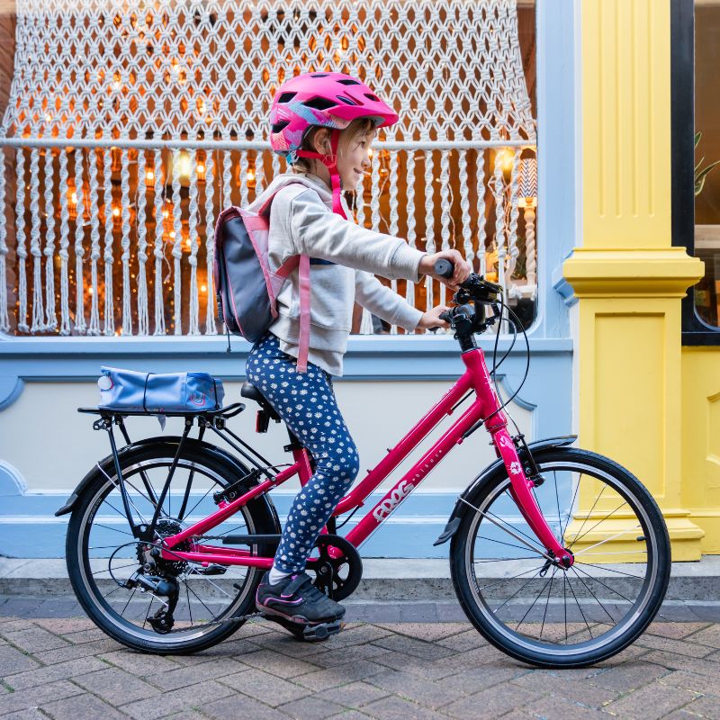 Child on a pink City 53 bicycle wearing a helmet and polka dot pants, with a decorative fence 